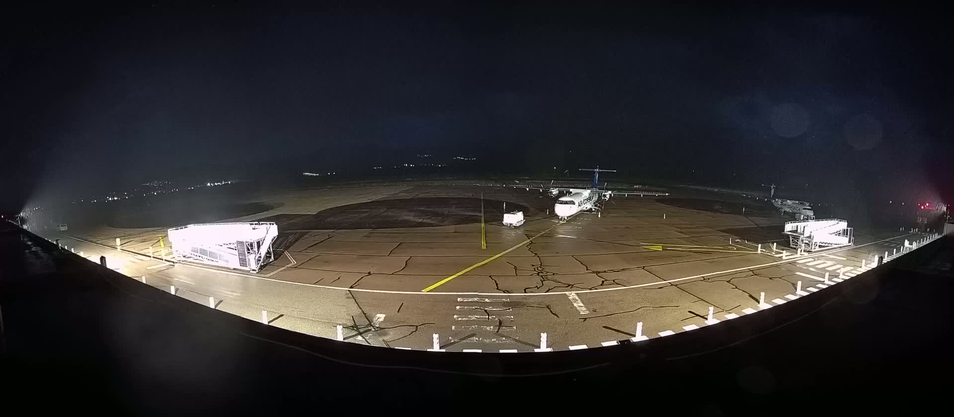 A wet airplane tarmac at night shows a small airplane parked near two jet bridges.