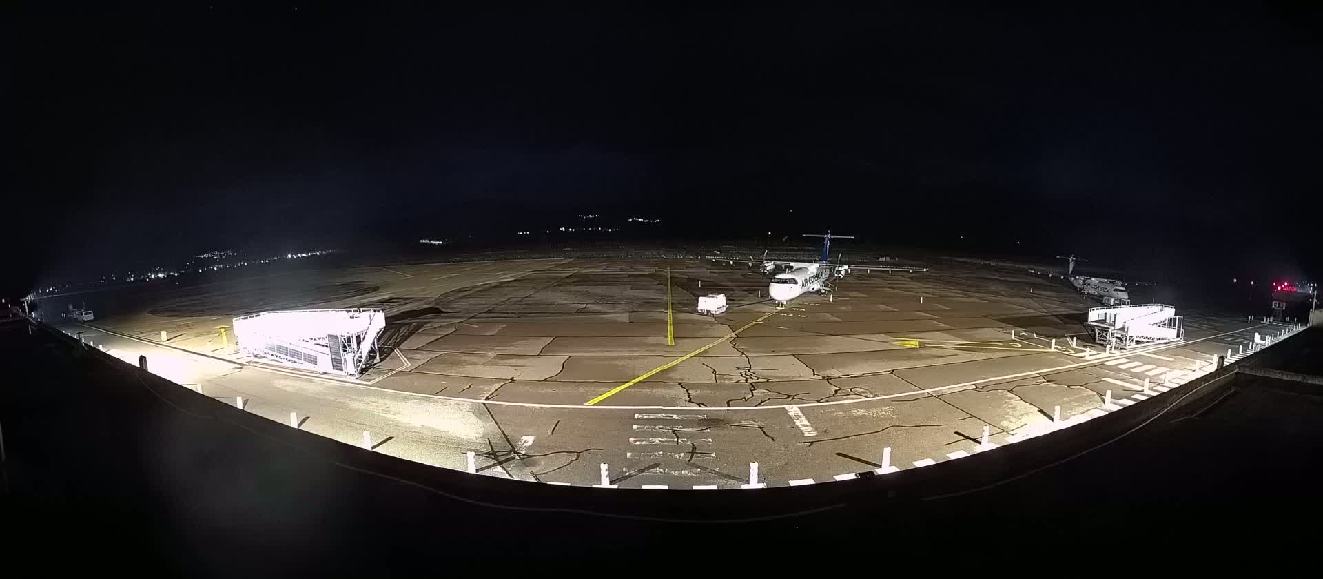 A nighttime, brightly lit airport tarmac shows a single airplane parked near two passenger boarding bridges.
