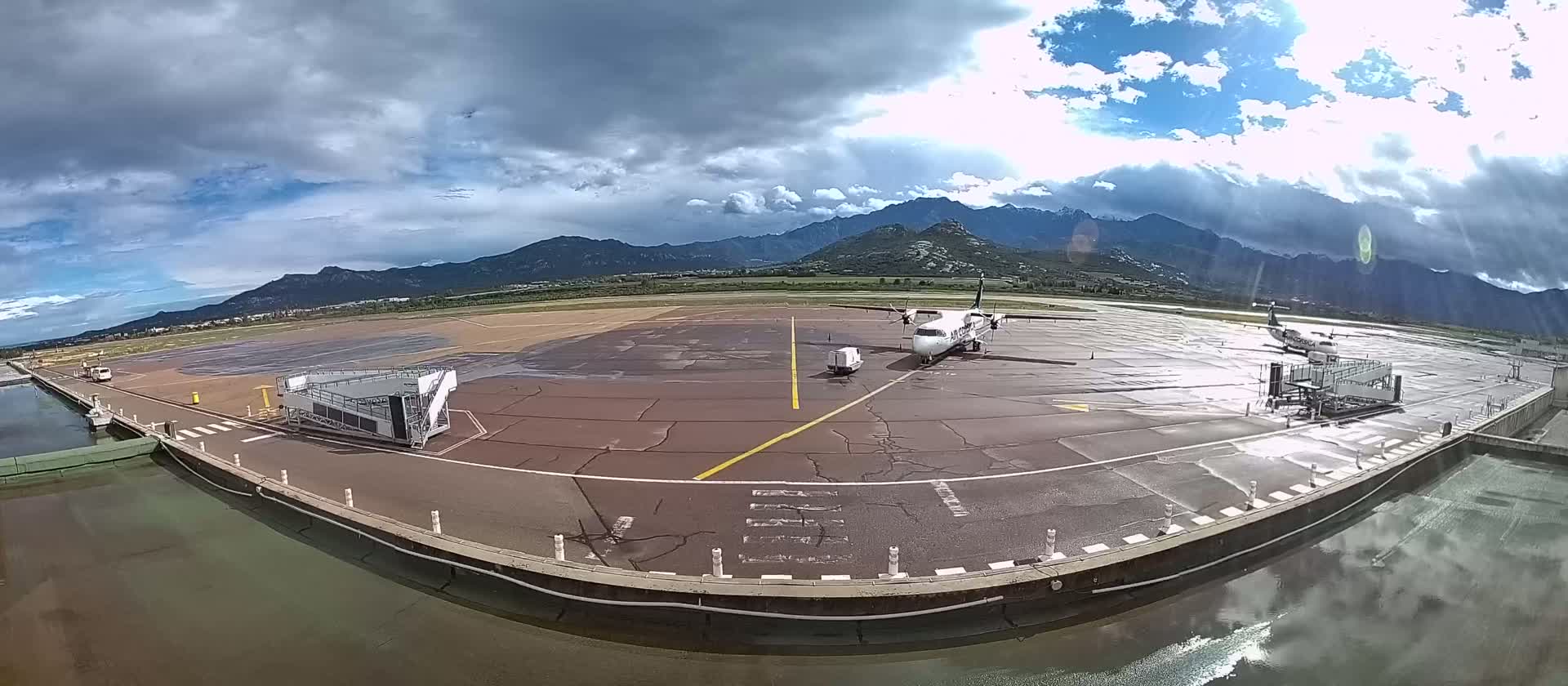 Two airplanes are parked at a gate on a wet tarmac, with mountains visible in the background under a partly cloudy sky.