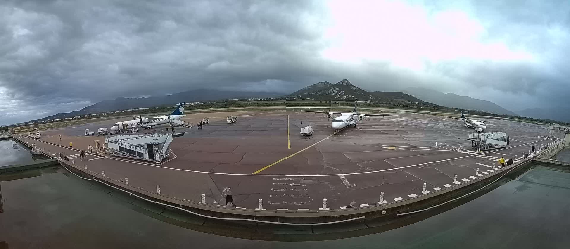 Three airplanes are parked at a tarmac near mountains under a cloudy sky.