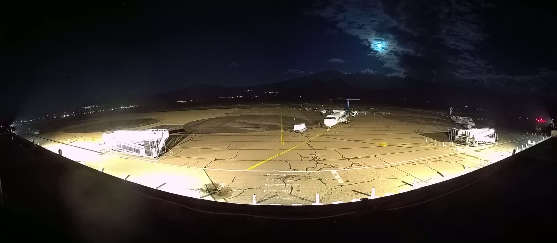 An airplane is parked on a tarmac at night under a partly cloudy moonlit sky, with jet bridges and ground support equipment nearby.
