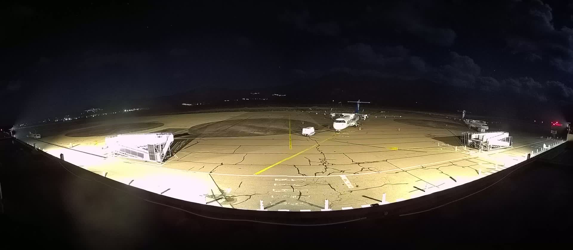 A nighttime, brightly lit airport tarmac shows a small passenger plane parked near two jet bridges under a partly cloudy sky.
