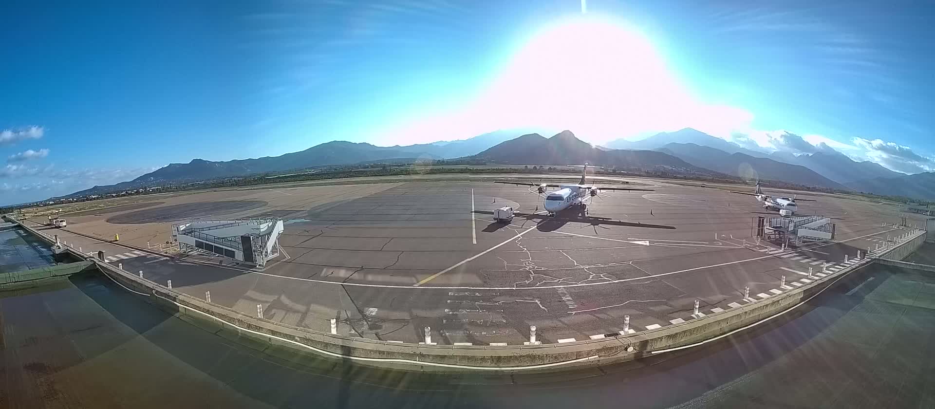 Two airplanes are parked at a tarmac with jet bridges attached, under a bright, sunny sky with mountains in the background.