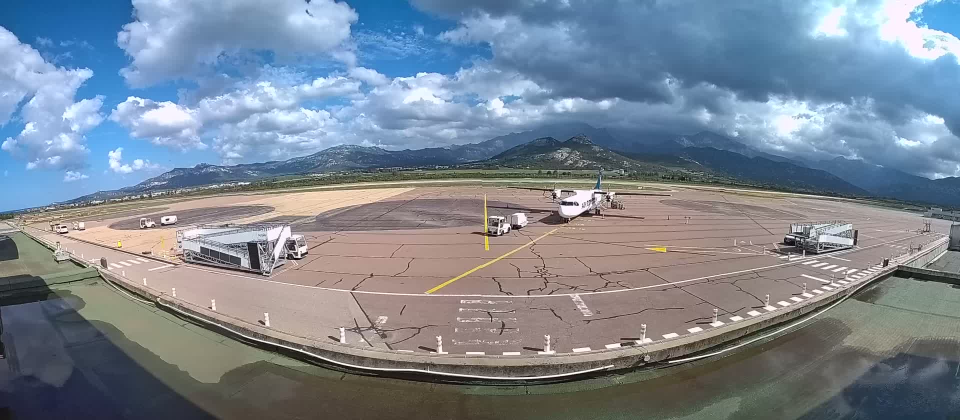 A propeller plane sits on a tarmac at an airport under a partly cloudy sky, with mountains visible in the distance.