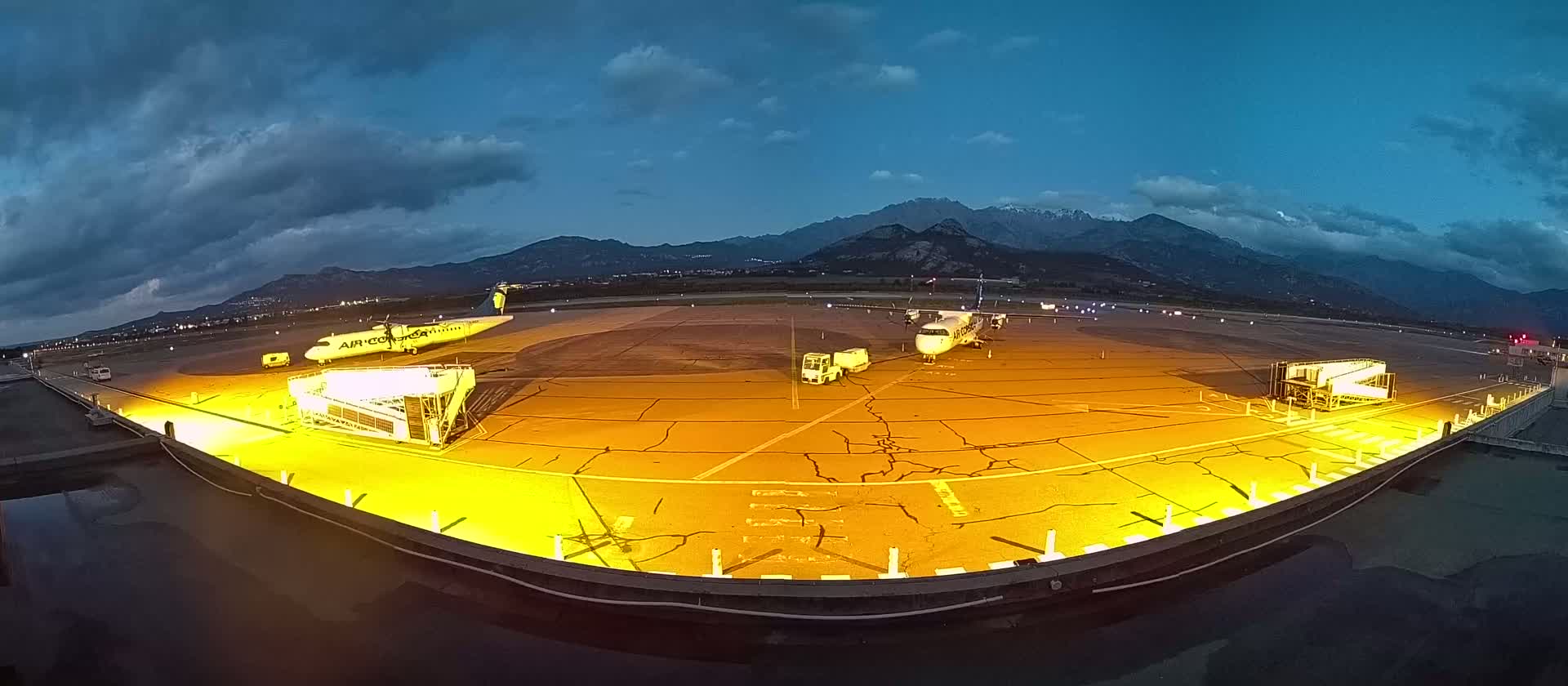 An airplane is parked on a brightly lit tarmac at dusk, with mountains in the background under a partly cloudy sky.