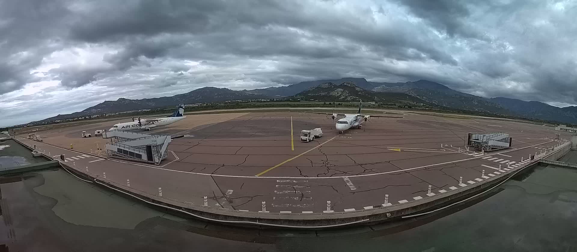 Two airplanes are parked at a tarmac with jet bridges, under a cloudy sky and mountains in the distance.