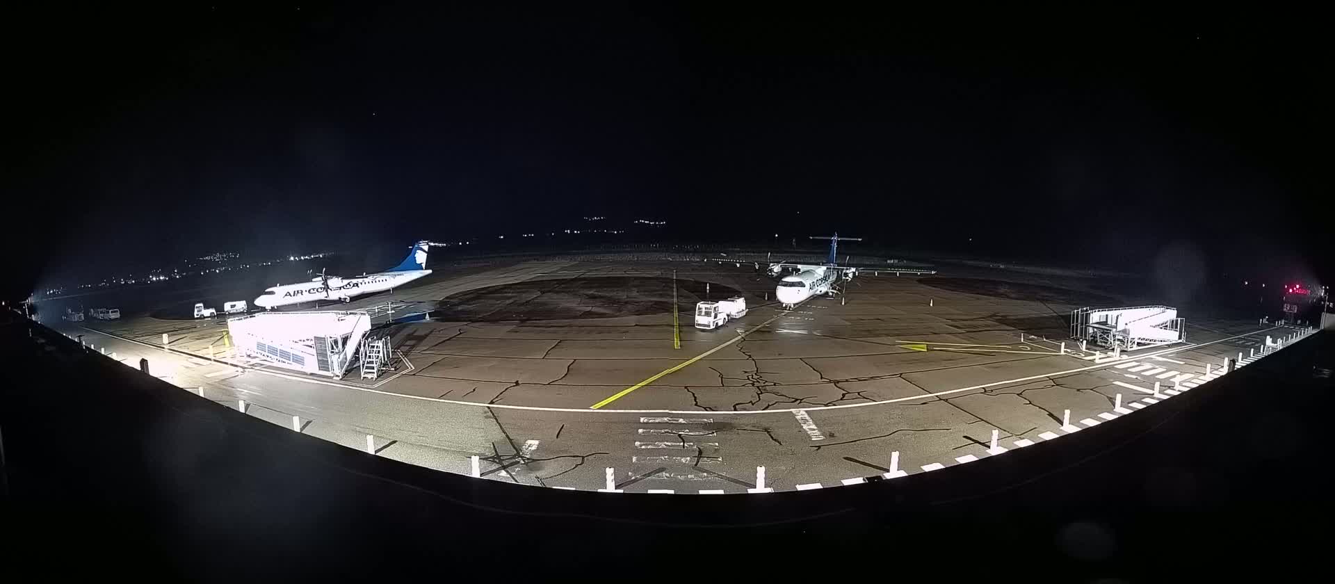 Two airplanes are parked on a dark tarmac at night, with one connected to a jet bridge and ground support vehicles nearby.
