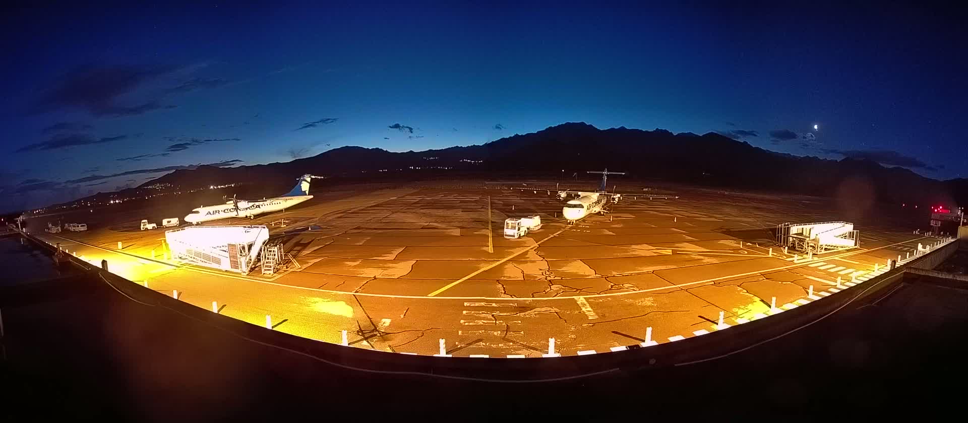 Two airplanes are parked on a brightly lit tarmac at night under a clear, dark blue sky with mountains in the distance.