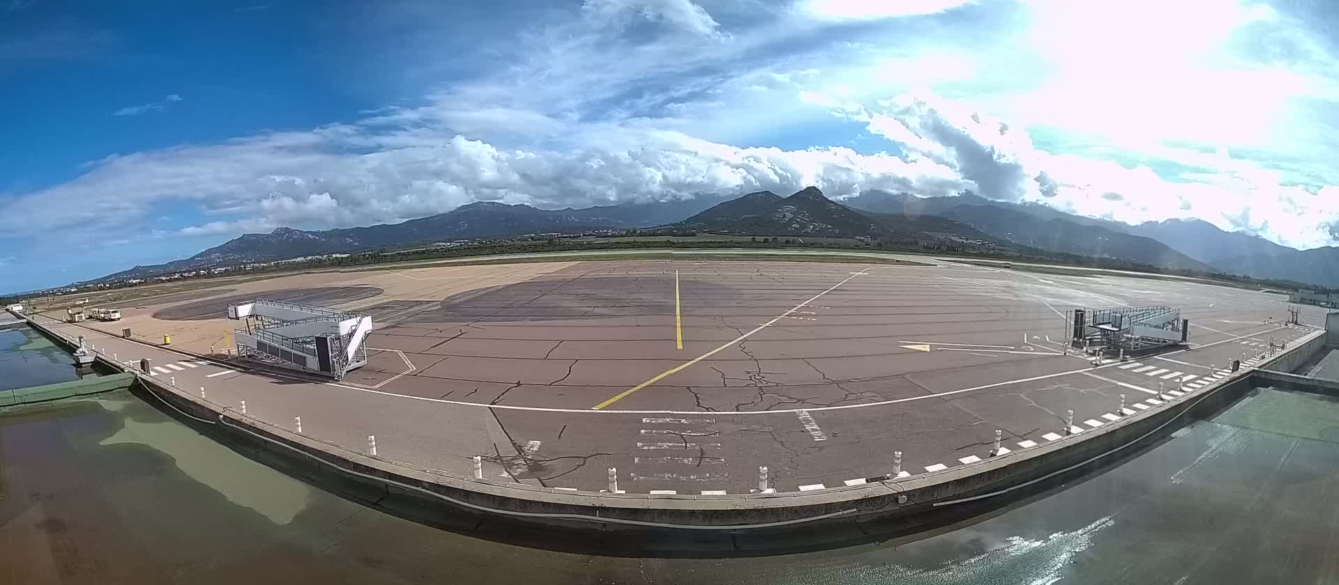 An airport tarmac with two jet bridges is visible, under a partly cloudy sky with mountains in the background.