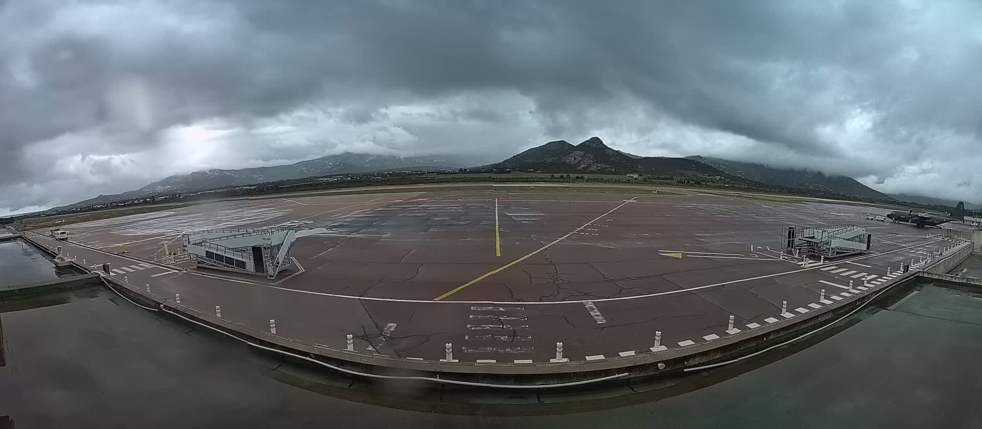 A wet airport tarmac under a cloudy sky, with mountains visible in the distance.