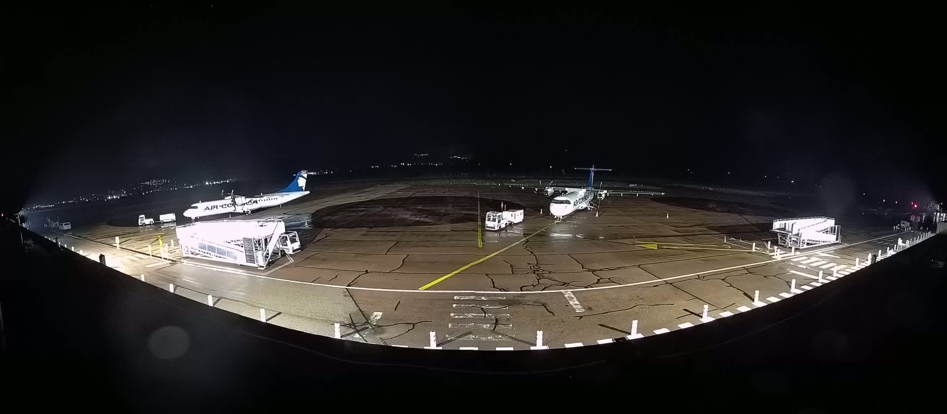 Two airplanes are parked on a wet tarmac at night, with ground support equipment nearby.