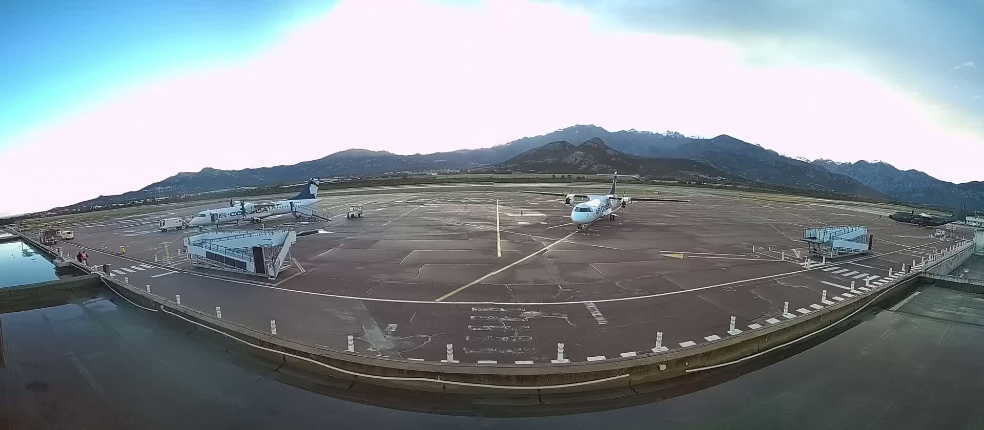 Two airplanes are parked on an airport tarmac, with mountains in the background under a mostly clear sky.