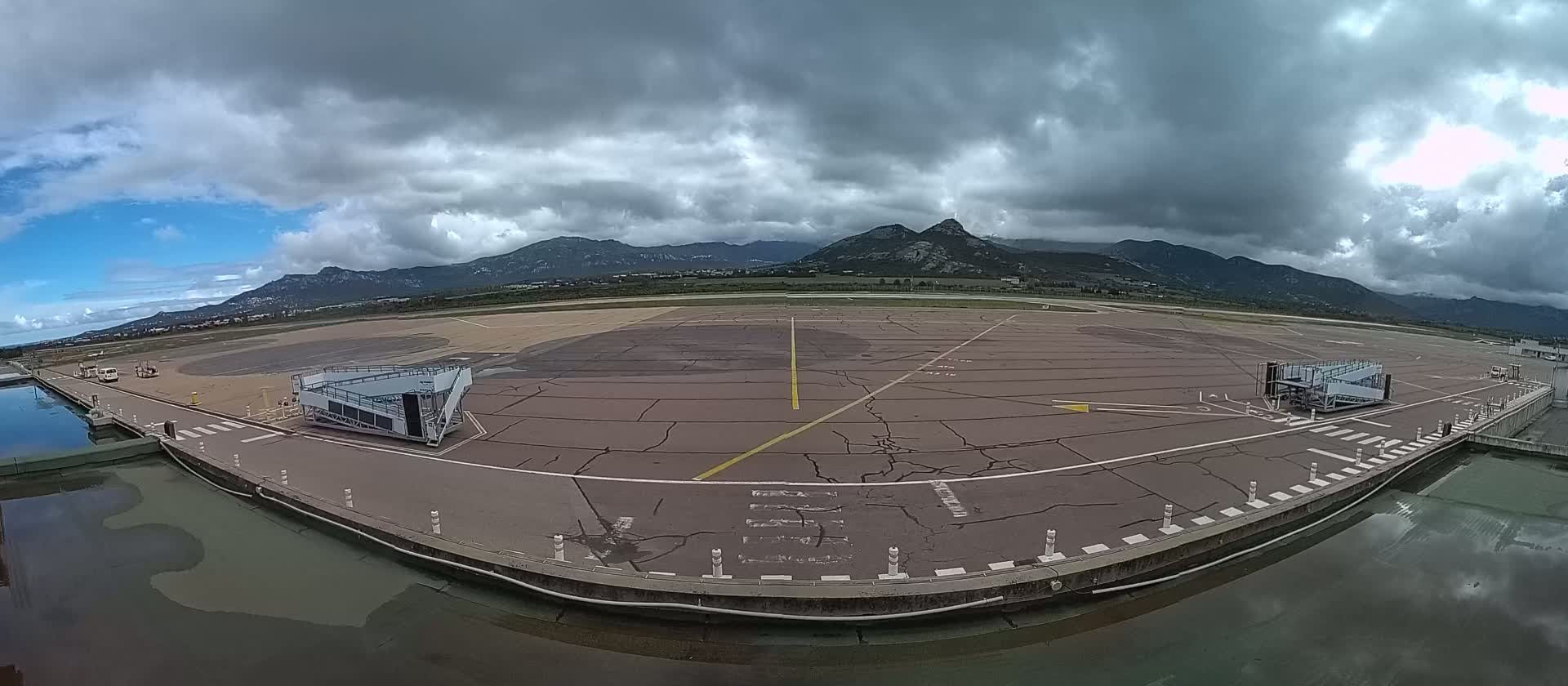 An airport tarmac with two jet bridges is visible under a cloudy sky, with mountains in the background.