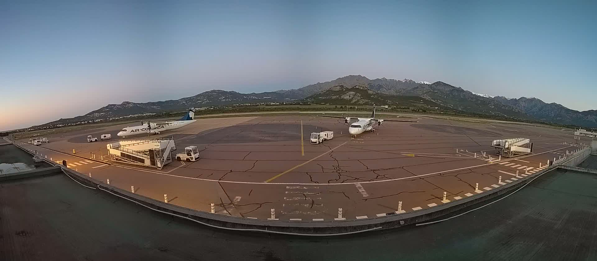 Two airplanes are parked on a tarmac at an airport, with jet bridges attached, under a twilight sky against a backdrop of mountains.
