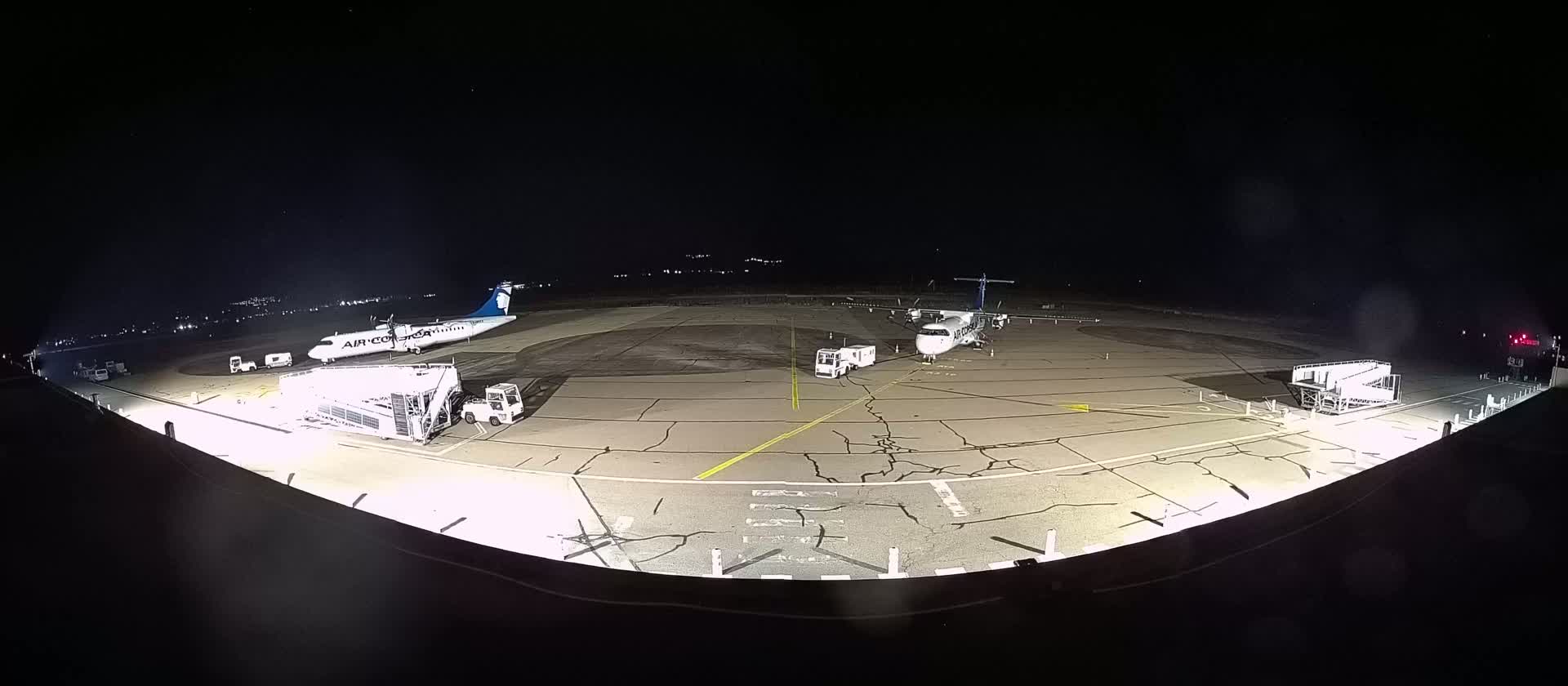 At night, two airplanes are parked on an airport tarmac illuminated by bright lights, with ground support equipment nearby.