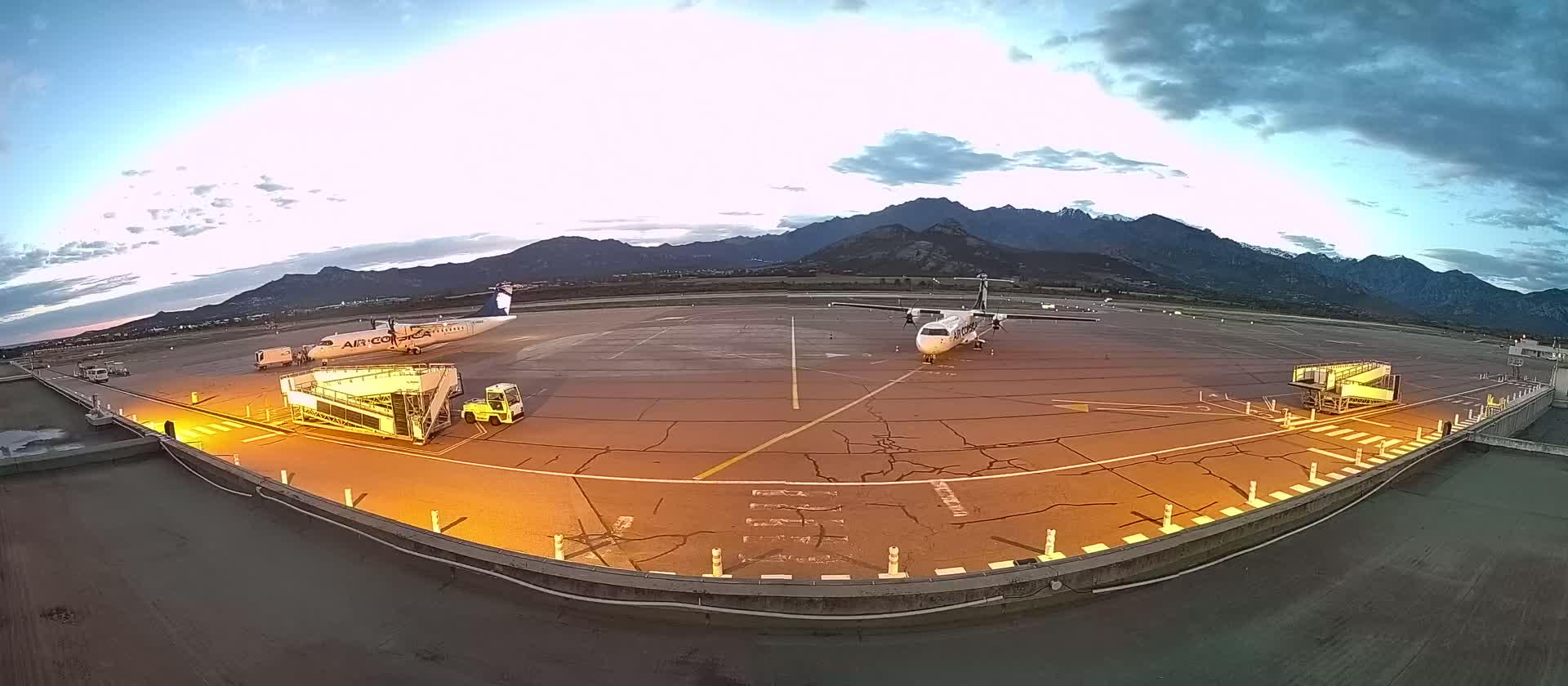 Two airplanes are parked at an airport tarmac at dusk, with mountains in the background under a partly cloudy sky.