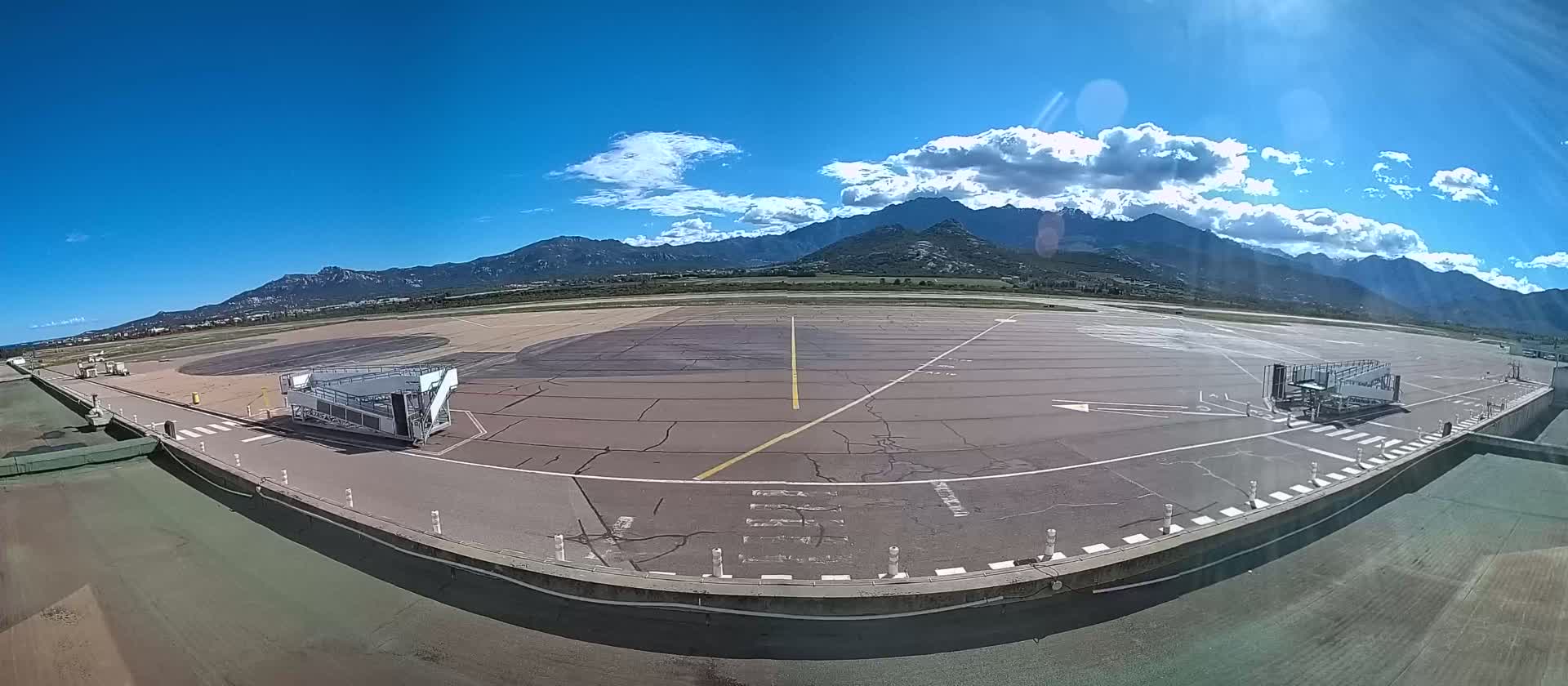 An airport tarmac with two jet bridges is visible under a mostly sunny sky with puffy clouds, mountains are in the background.