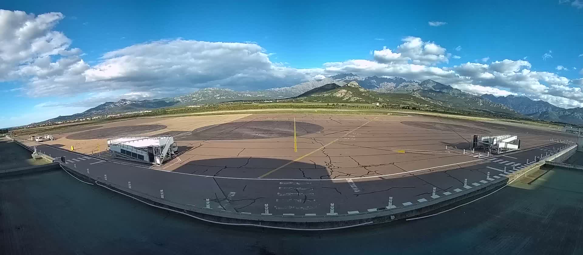 An airport tarmac with two passenger boarding bridges is visible, under a partly cloudy sky with mountains in the background.