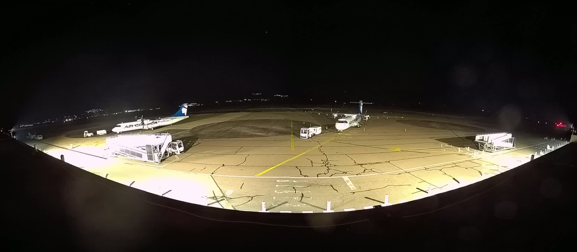 At night, two airplanes are parked on a tarmac illuminated by bright lights, with ground support equipment nearby.