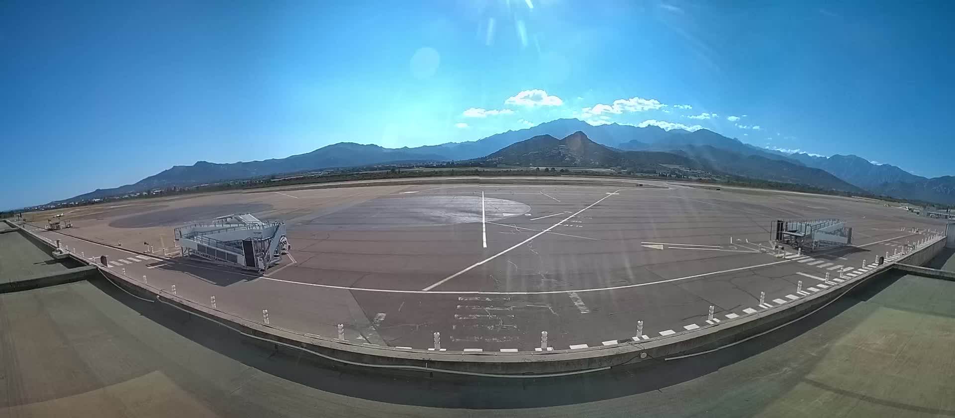 A tarmac area at an airport, with two jet bridges and mountains in the background under a clear, sunny sky.