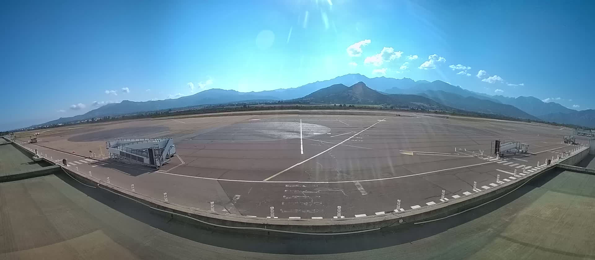 A mostly empty airport tarmac with a jet bridge, under a bright, sunny sky, and mountains in the background.