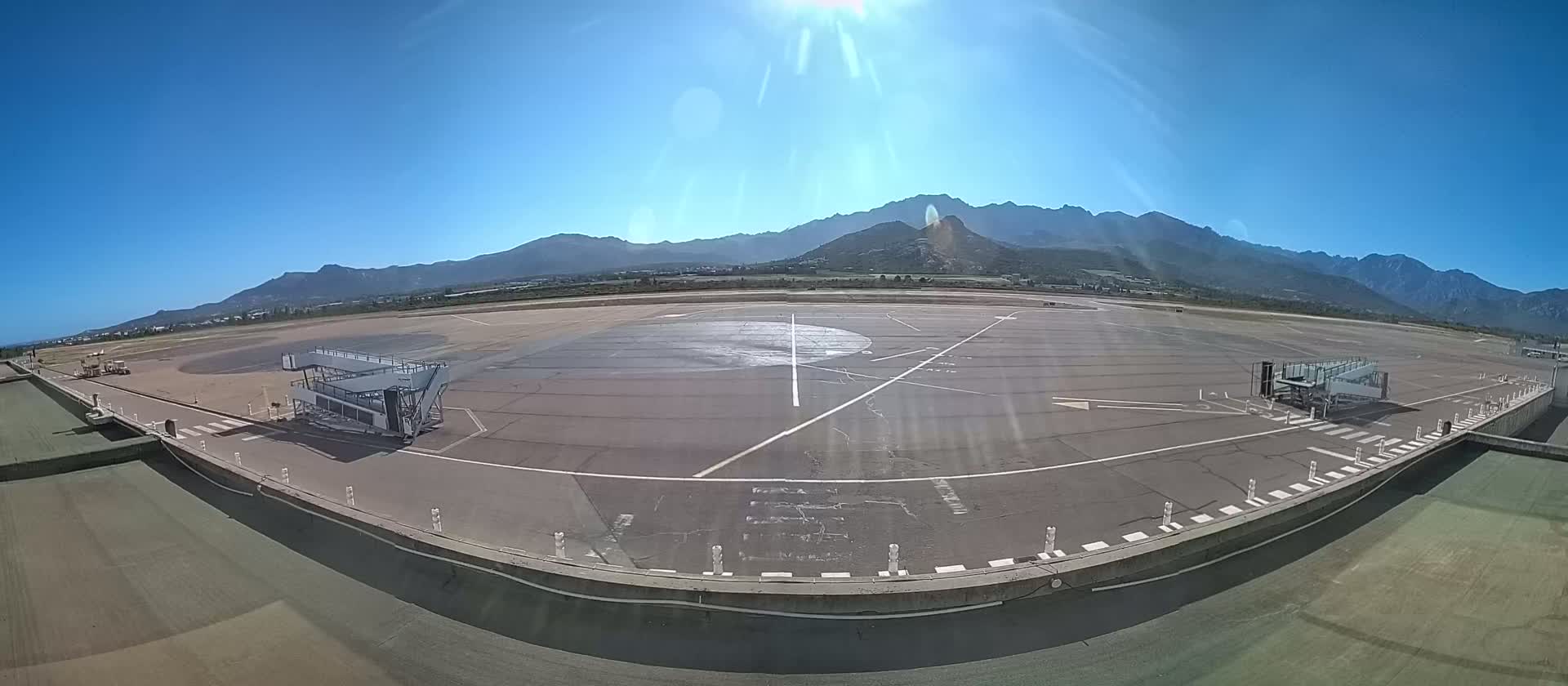 A sunny airport tarmac with two jet bridges is visible, backed by a mountain range under a clear blue sky.