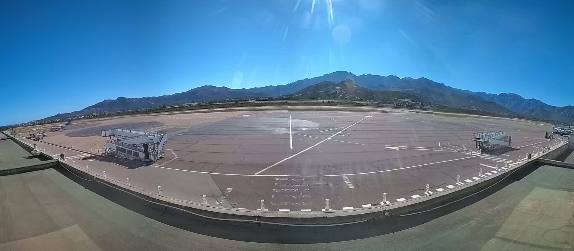 An airport tarmac with two jet bridges is visible under a clear blue sky, with mountains in the background.