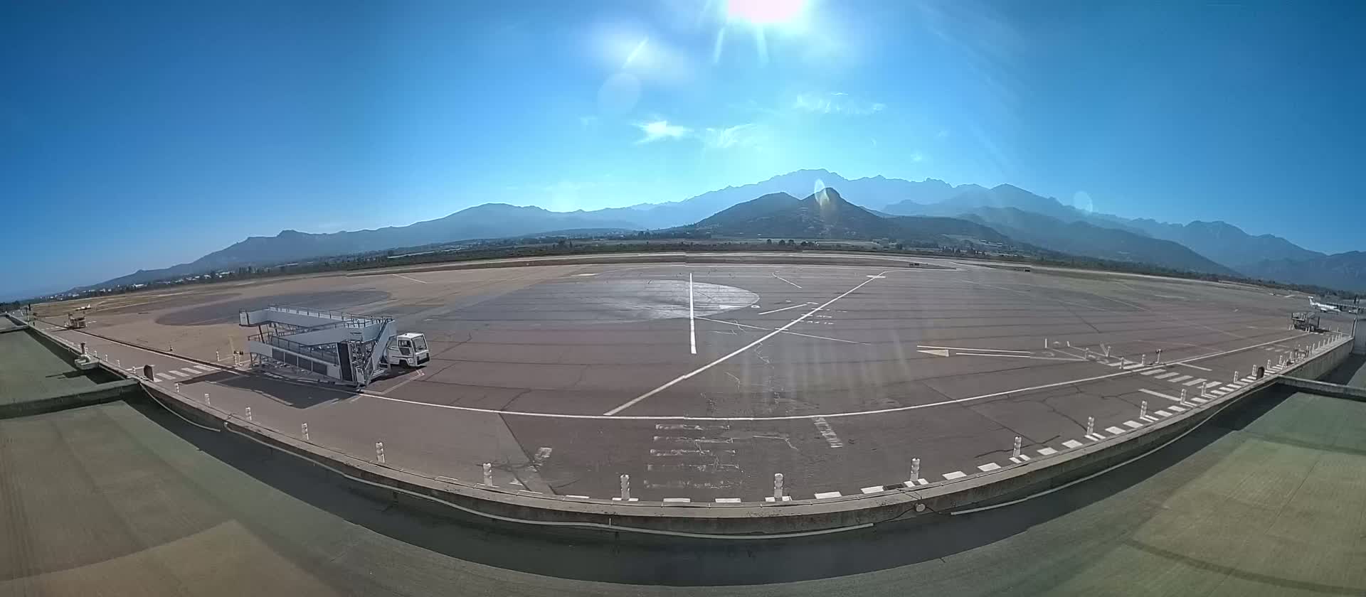 A jet bridge and support vehicle sit on a tarmac at an airport under a bright, sunny sky with mountains in the background.