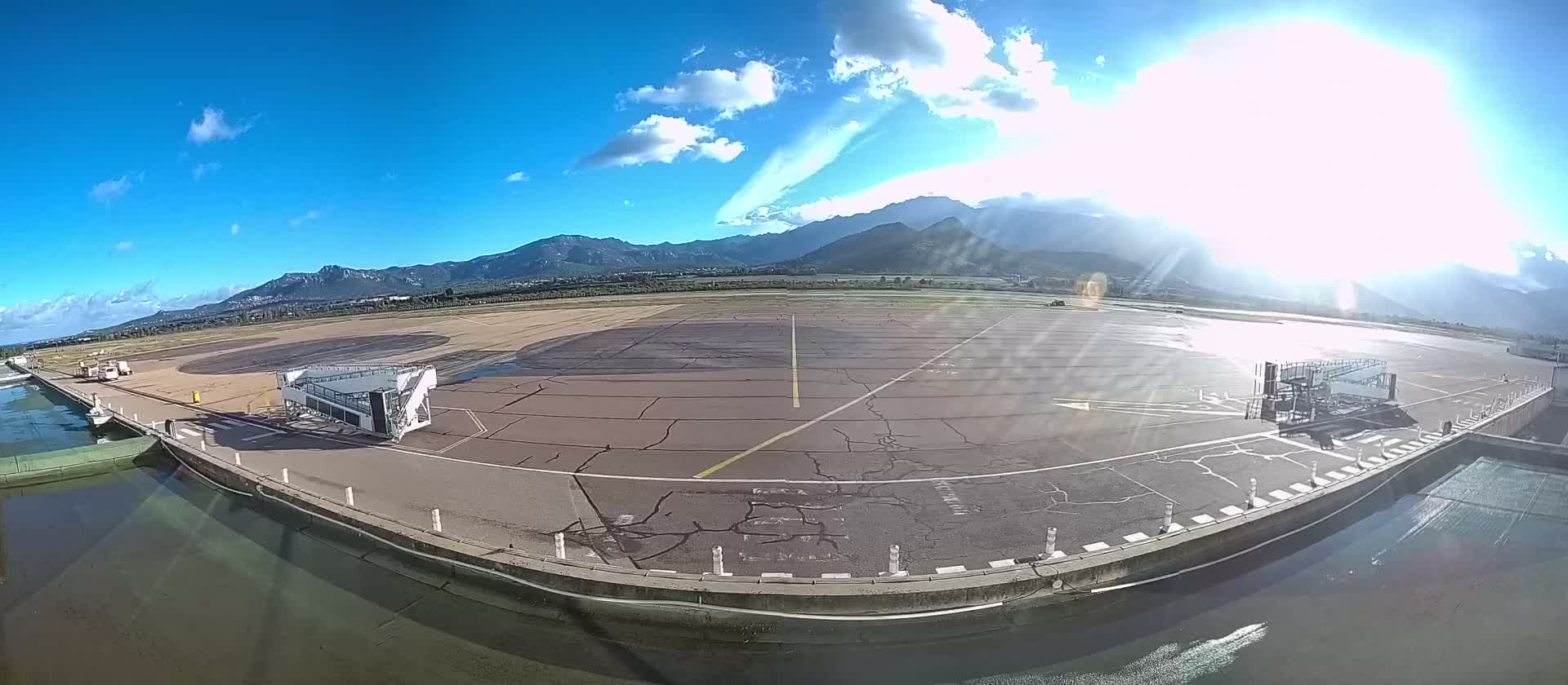 An expansive airport tarmac with two mobile jet bridges and a foreground waterway stretches towards distant mountains under a bright, sunny blue sky with scattered white clouds.