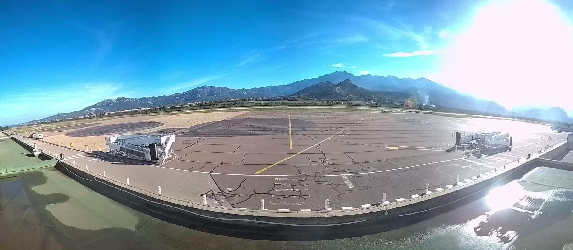 On a bright, sunny day with a clear blue sky, an airport apron with cracked asphalt and markings, featuring two mobile passenger staircases, a small white vehicle, and distant mountains, is seen above a reflective puddle in the foreground.