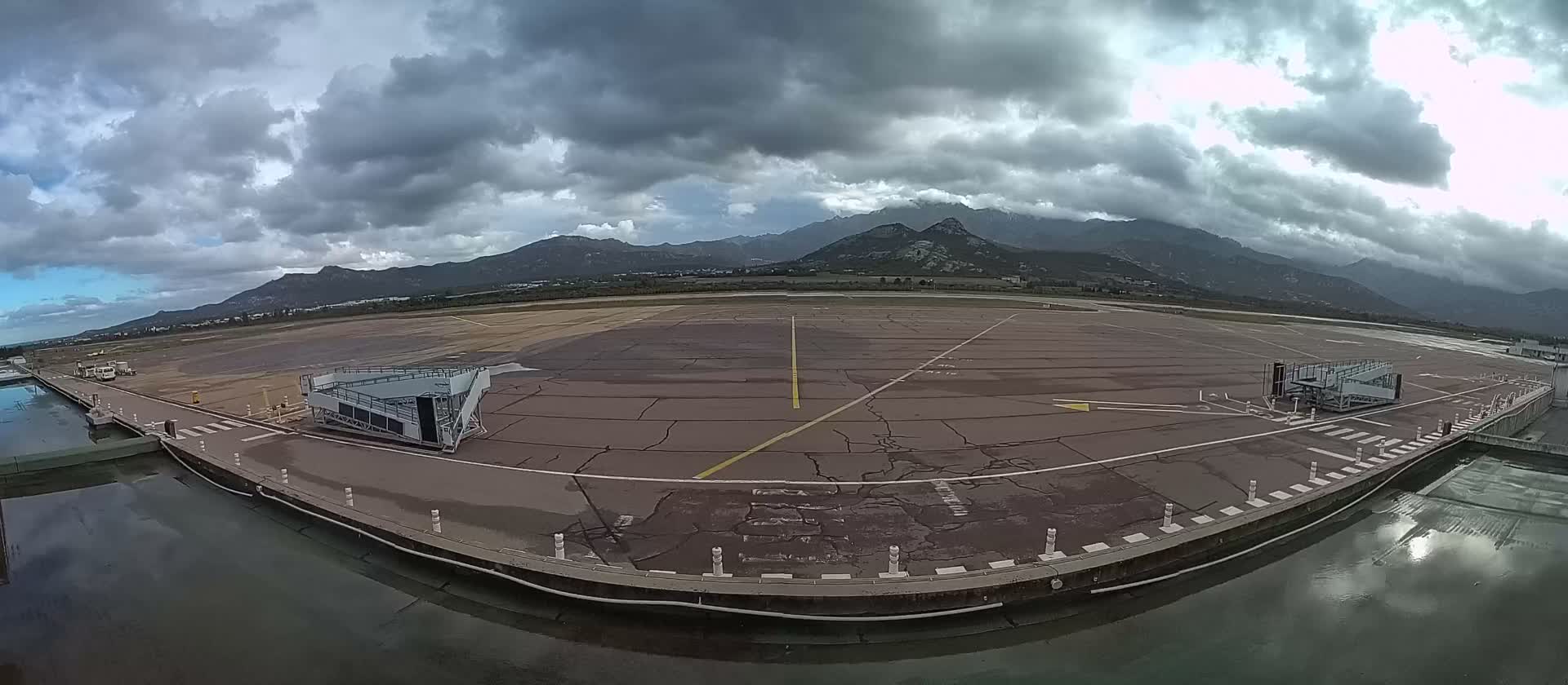 An airport tarmac with wet patches and mobile boarding stairs is depicted under a heavily overcast sky with mountains in the background, bordered by a calm reflecting pool in the foreground.