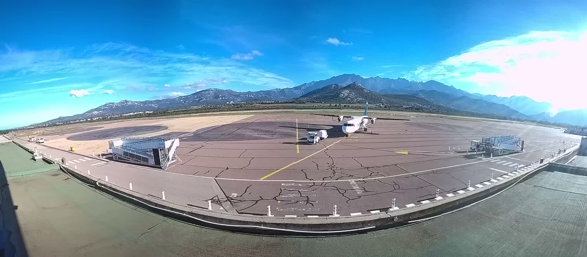 An airport tarmac with wet patches and mobile boarding stairs is depicted under a heavily overcast sky with mountains in the background, bordered by a calm reflecting pool in the foreground.