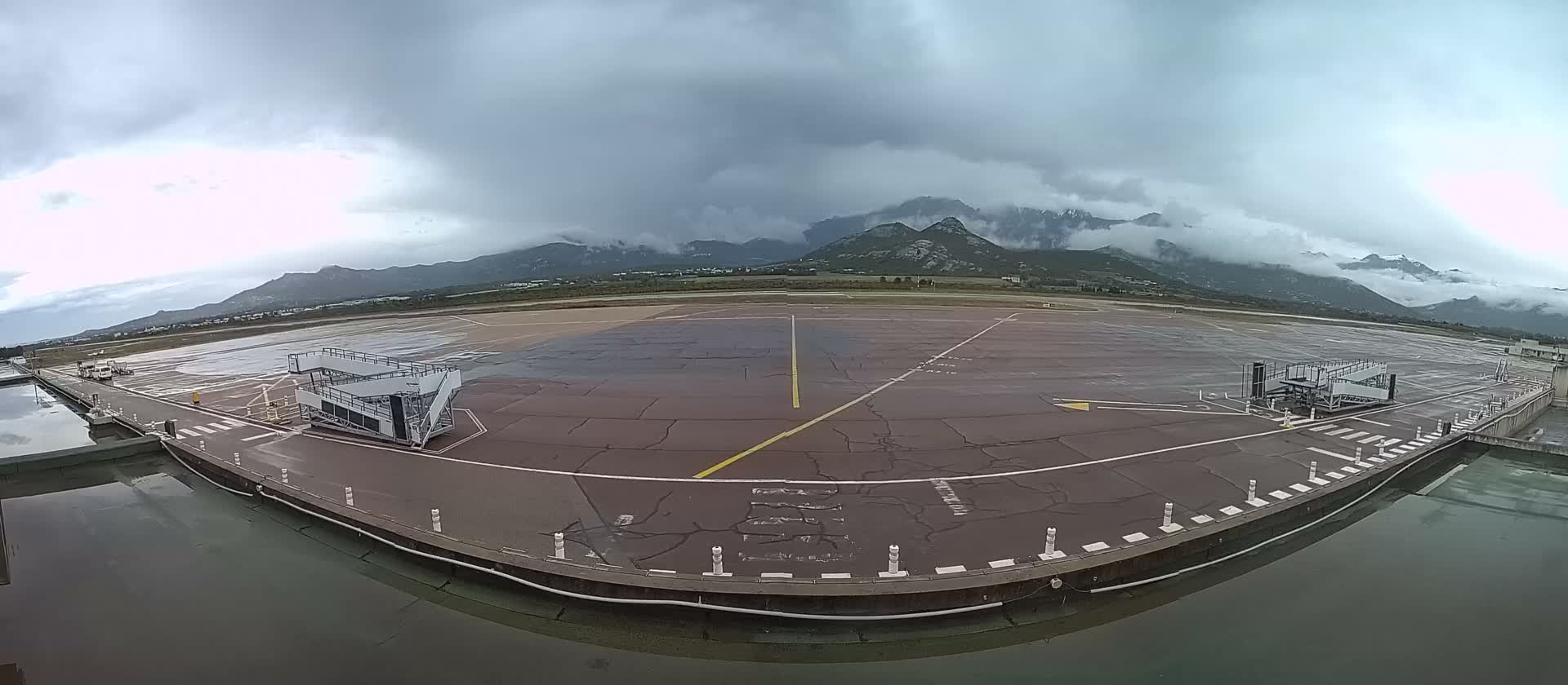 An airport tarmac with wet patches and mobile boarding stairs is depicted under a heavily overcast sky with mountains in the background, bordered by a calm reflecting pool in the foreground.
