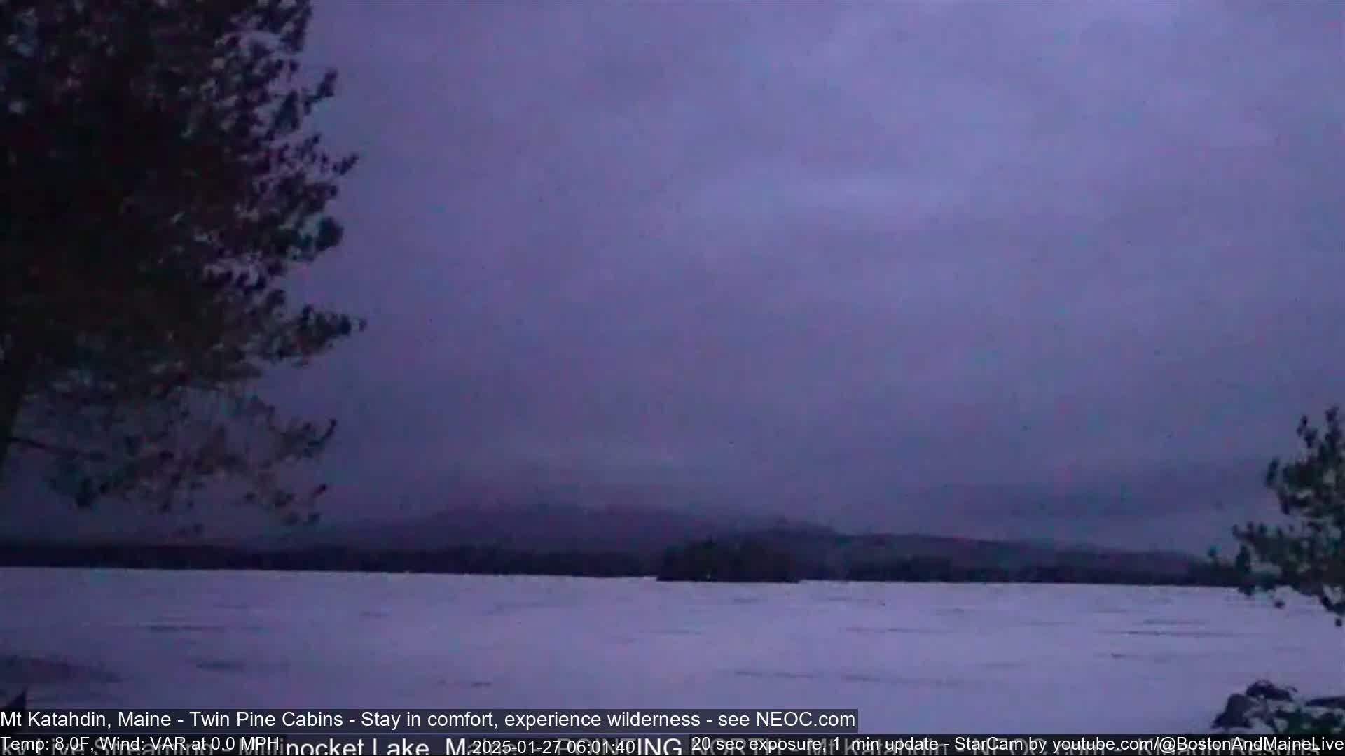 Here is a description of the image: A twilight view shows a snow-covered lake with dark trees silhouetted against a dusky purple sky.