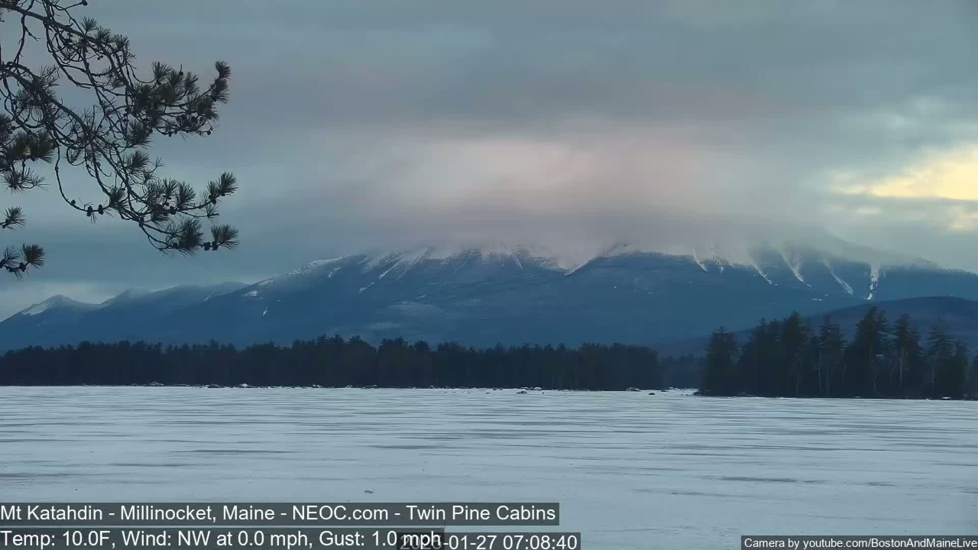 A snow-covered frozen lake stretches out before a line of snow-capped mountains under a cloudy sky.