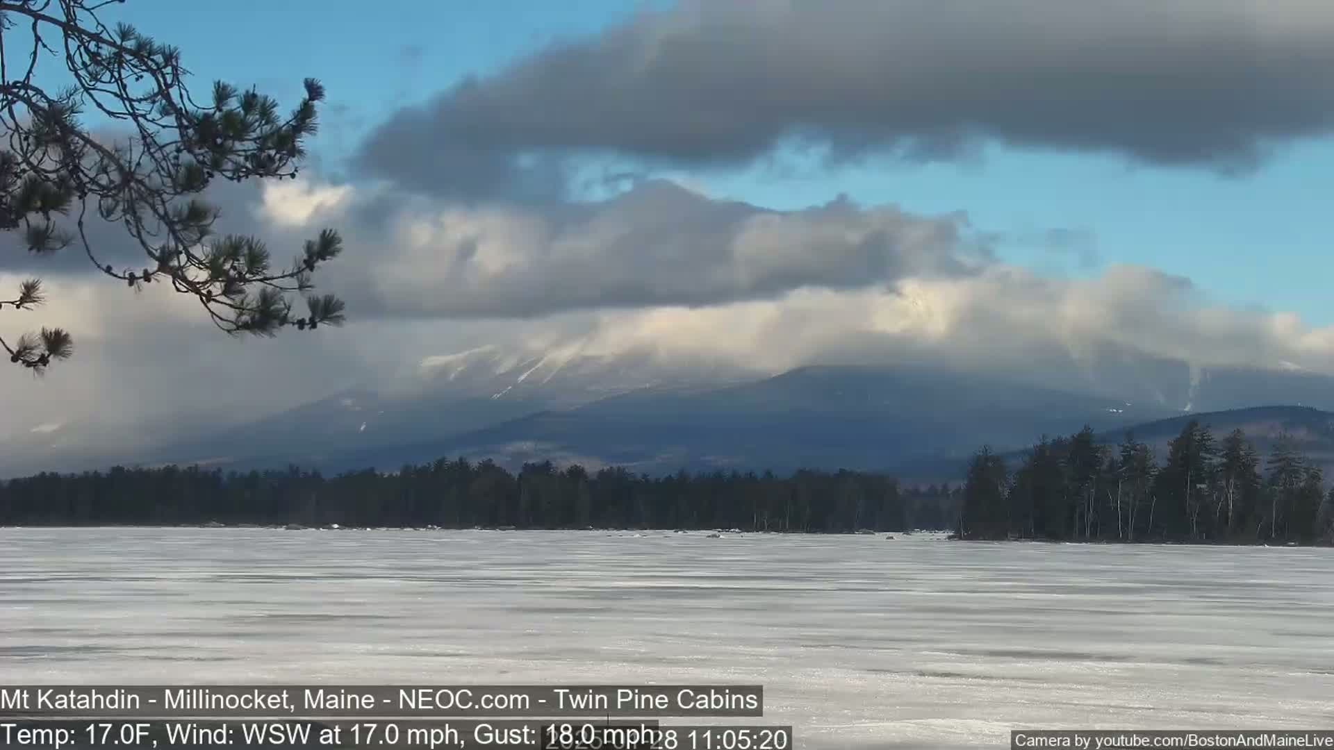 A partly cloudy, windy day with a frozen lake in the foreground and snow-capped mountains in the background.