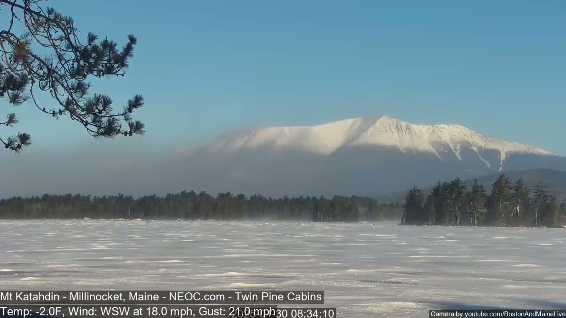 A snow-covered mountain is visible in the distance across a frozen lake under a clear, bright sky, with a moderate wind blowing.