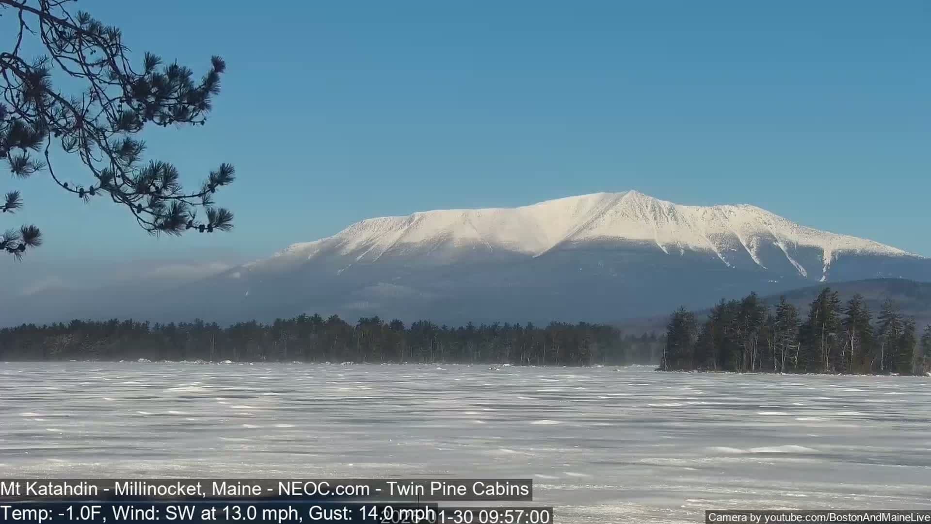 A snow-covered mountain is seen in the distance across a frozen lake under a clear blue sky, with a southwest wind blowing at 13 mph, gusting to 14 mph.