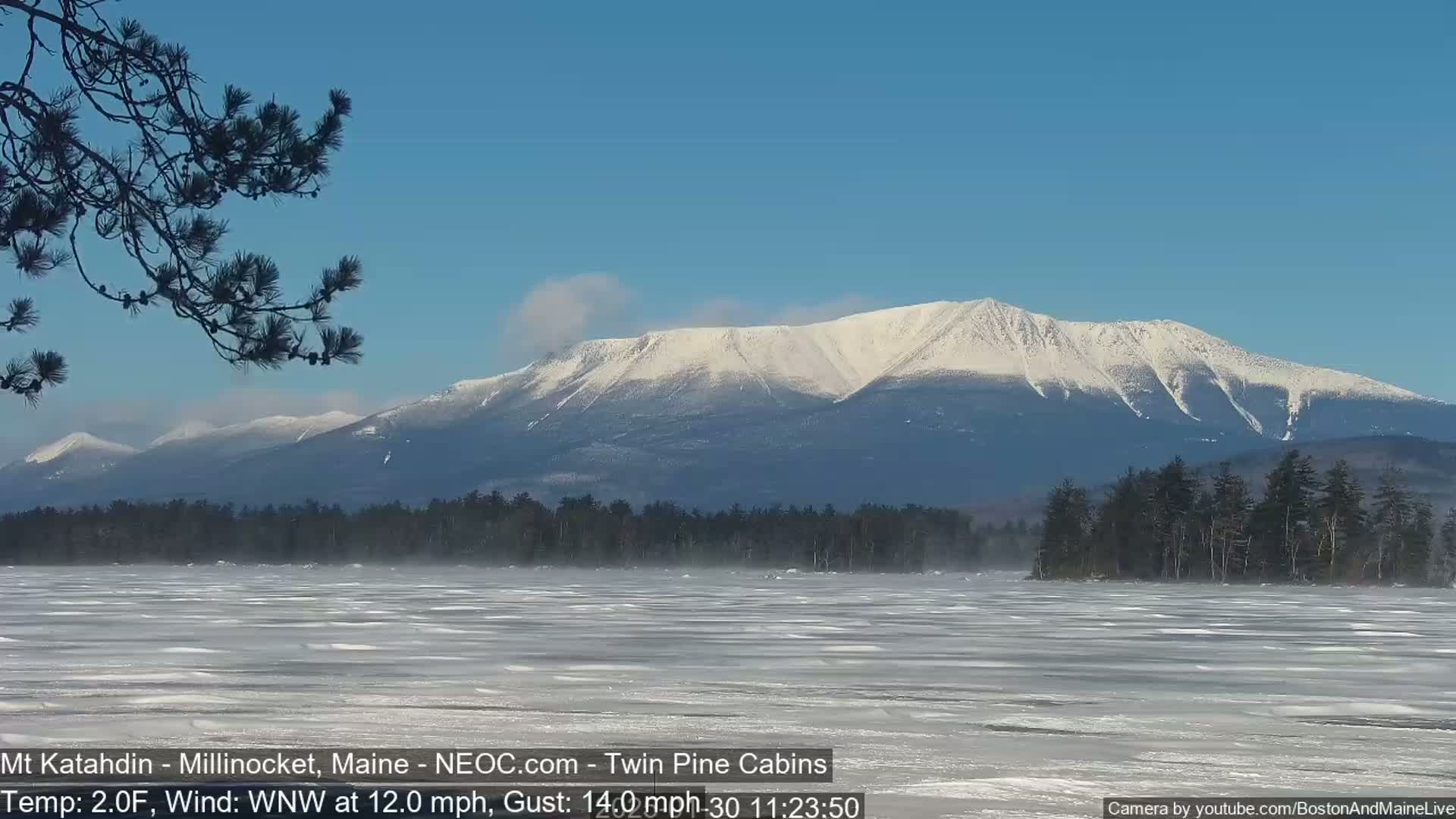 A snow-covered mountain range is visible in the distance across a frozen lake under a clear blue sky, with a wind blowing across the ice.