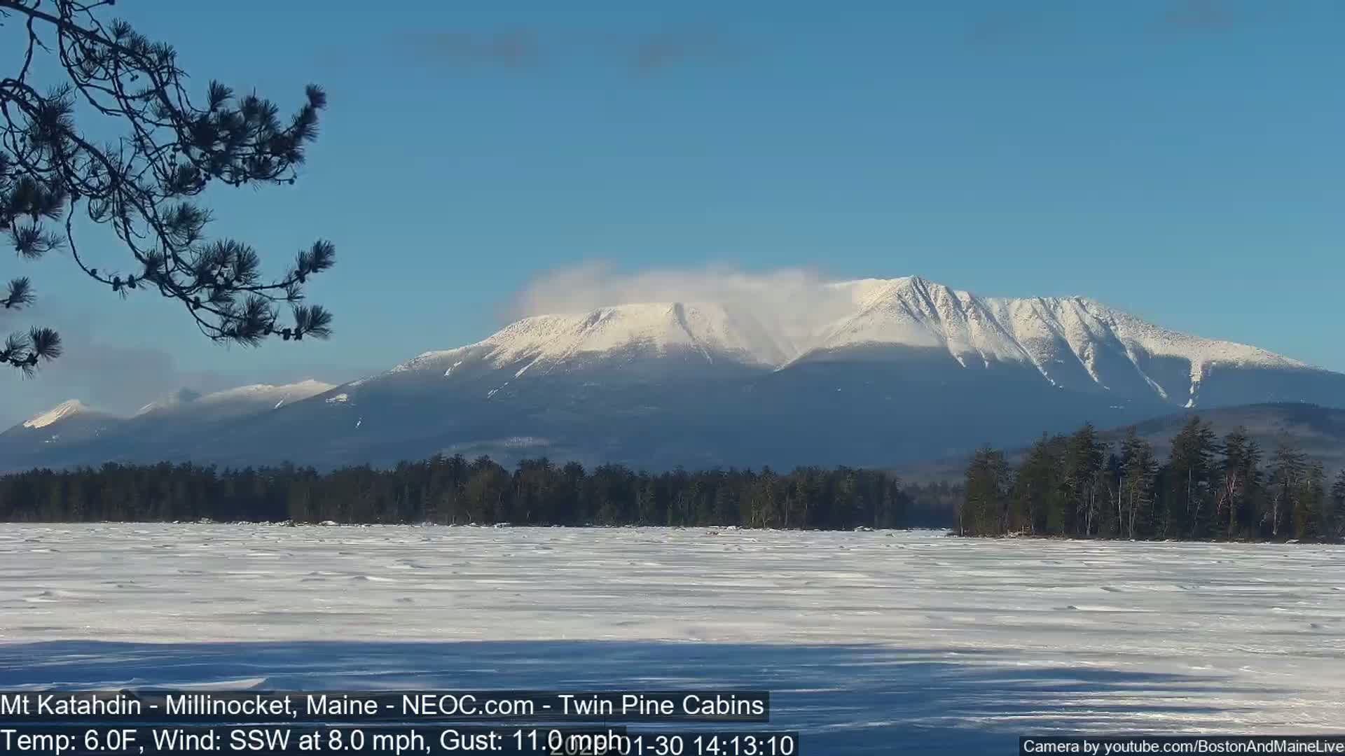 A snow-covered mountain range is visible in the distance across a frozen lake under a clear blue sky, with a light wind blowing.