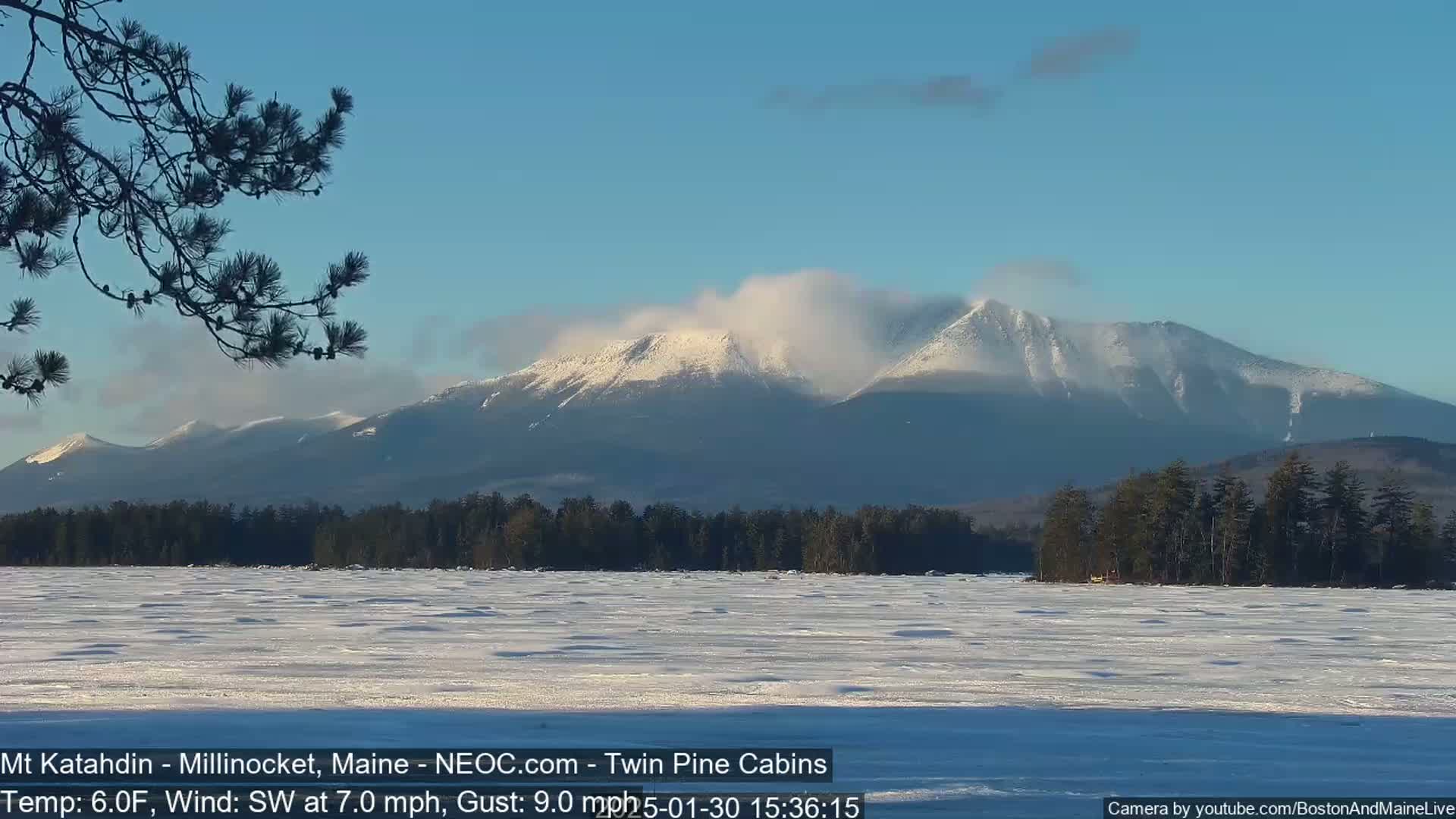 A snow-covered mountain range is visible in the distance across a frozen lake under a clear blue sky with a light southwest wind.