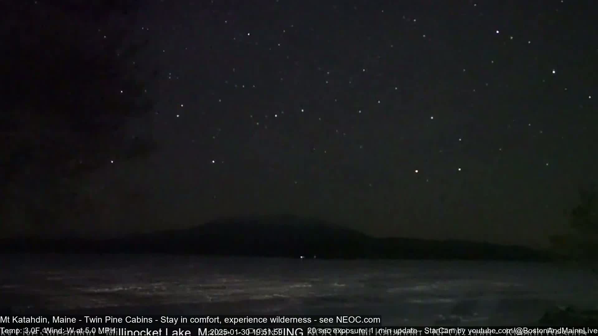 A dark, starlit night scene shows a calm body of water in front of a silhouetted mountain range.