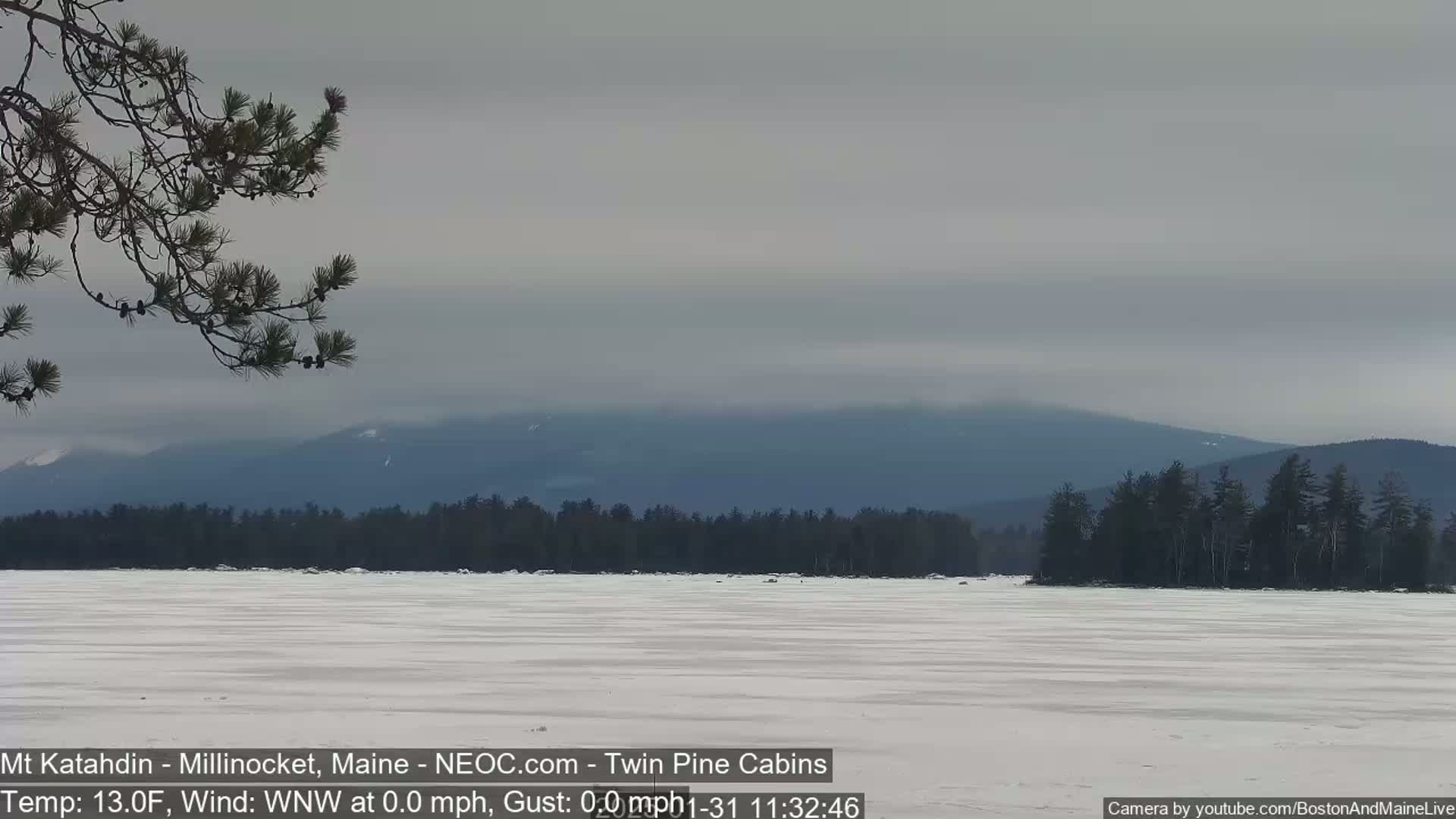 A snow-covered frozen lake stretches out before a line of dark evergreen trees, with a hazy mountain range visible in the distance under a mostly cloudy sky.