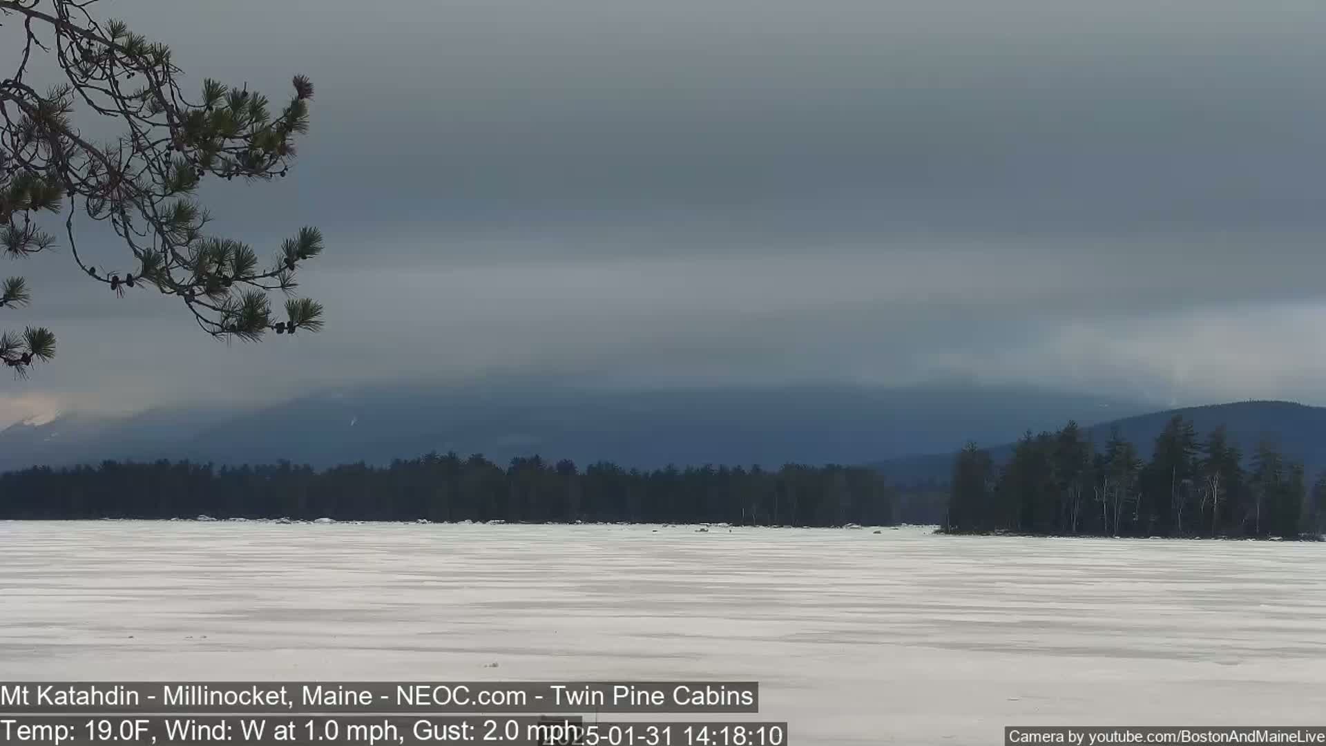 A snow-covered lake stretches out before a dark, overcast sky and a line of evergreen trees, with distant mountains visible in the background.