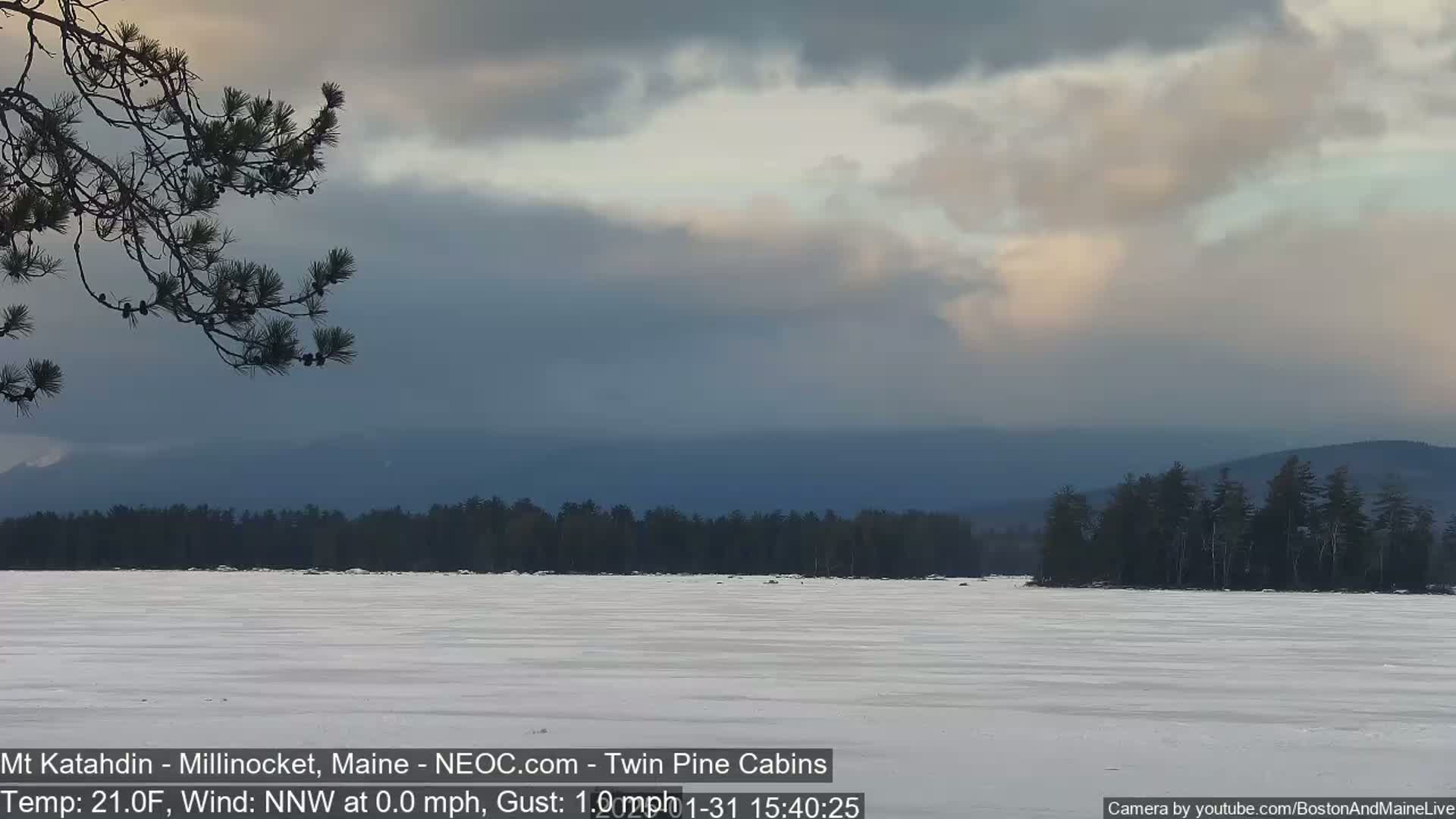 A snow-covered frozen lake stretches to a dark, tree-lined shore beneath a cloudy, pale sky, with distant mountains visible.