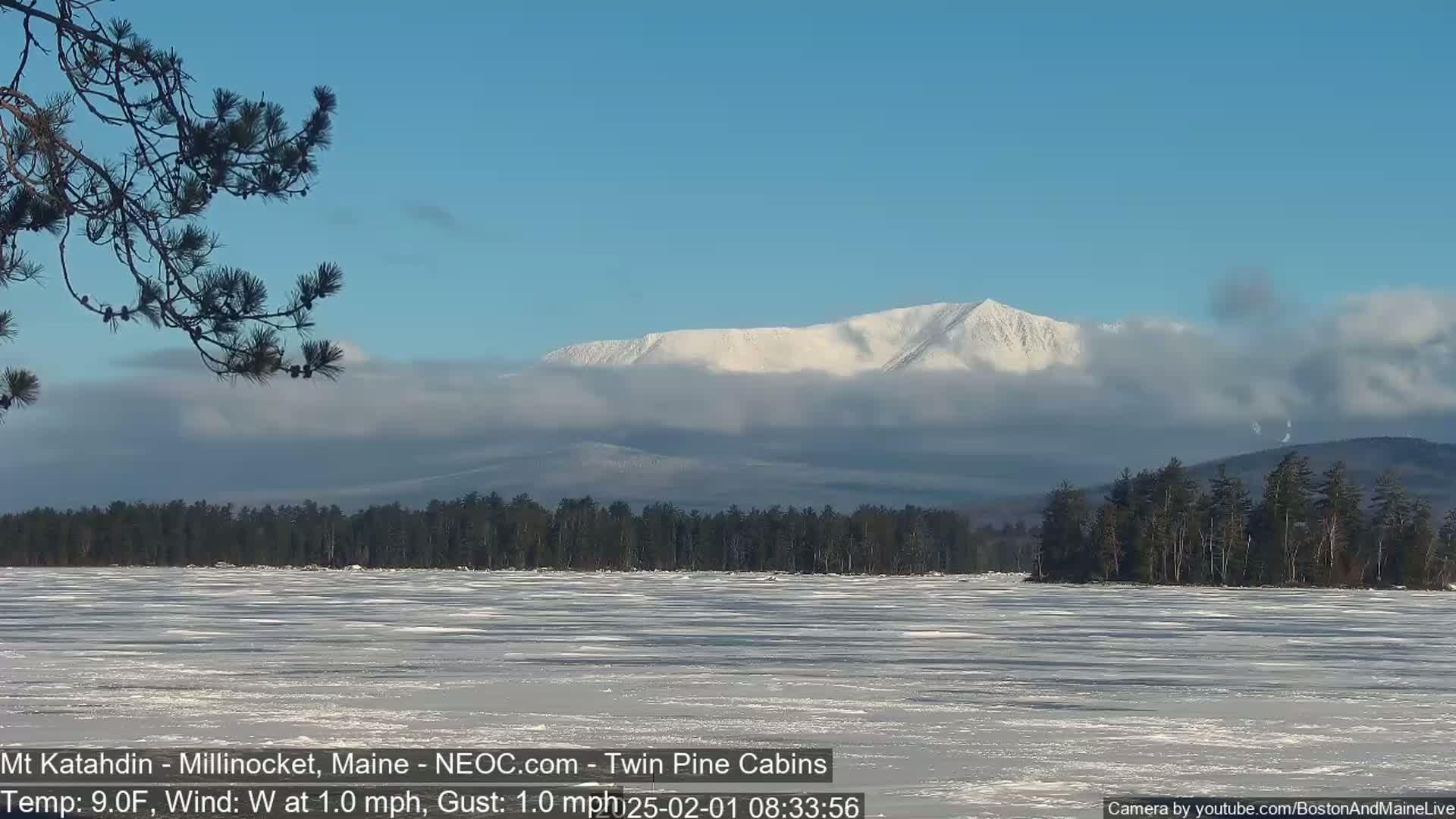 A partially frozen lake stretches to a snow-covered mountain partially obscured by clouds under a clear blue sky.