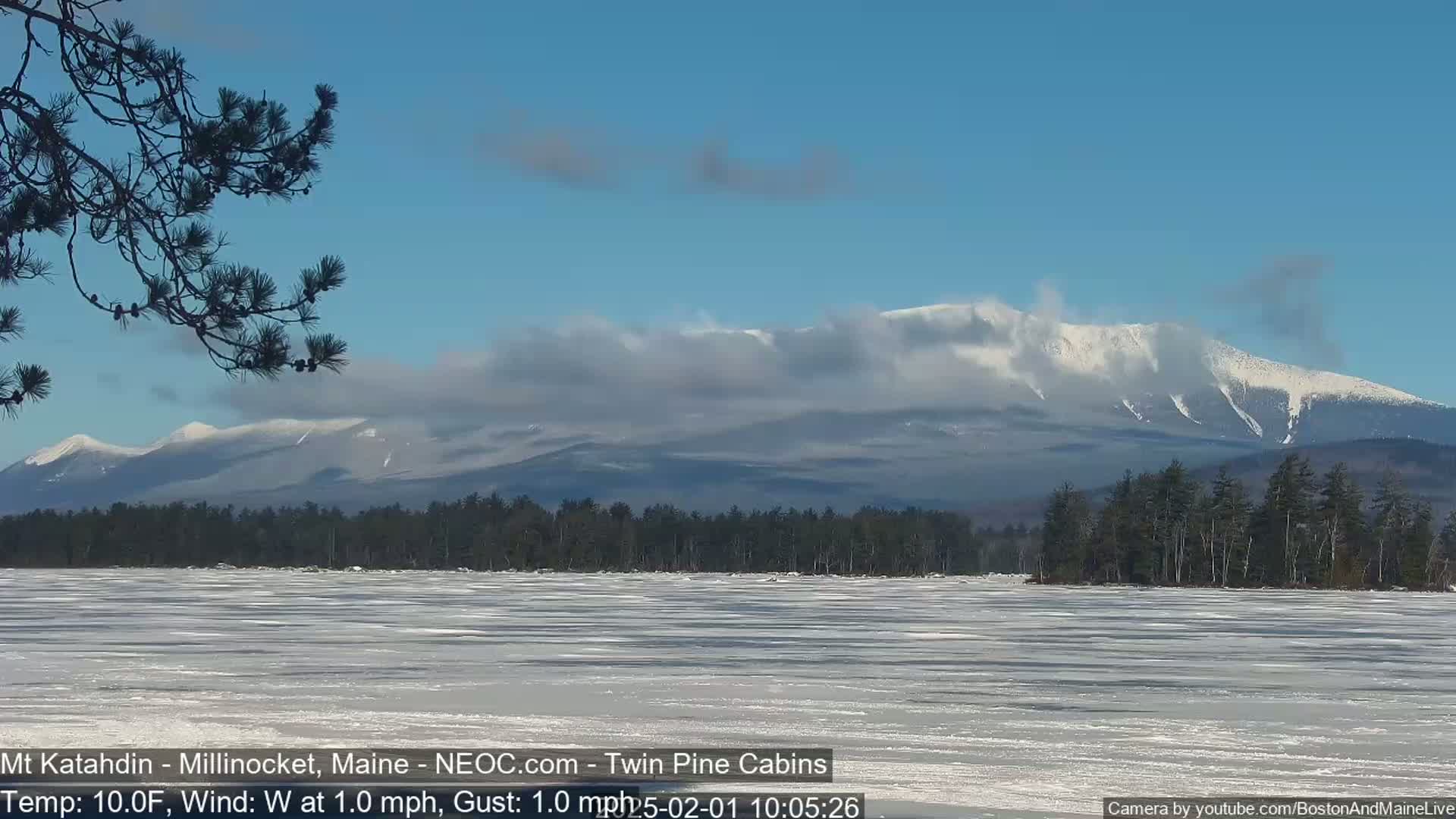 A partially frozen lake stretches out before a snow-covered mountain range under a mostly clear, bright sky.