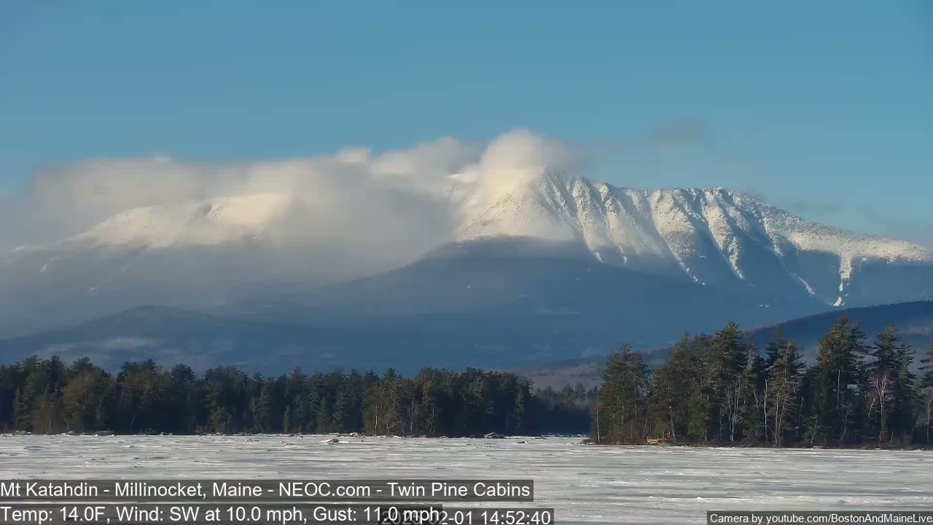 A snow-covered mountain range is visible under a mostly clear sky, with a frozen lake and dark evergreen trees in the foreground, and a southwest wind blowing at 10 mph with gusts up to 11 mph.