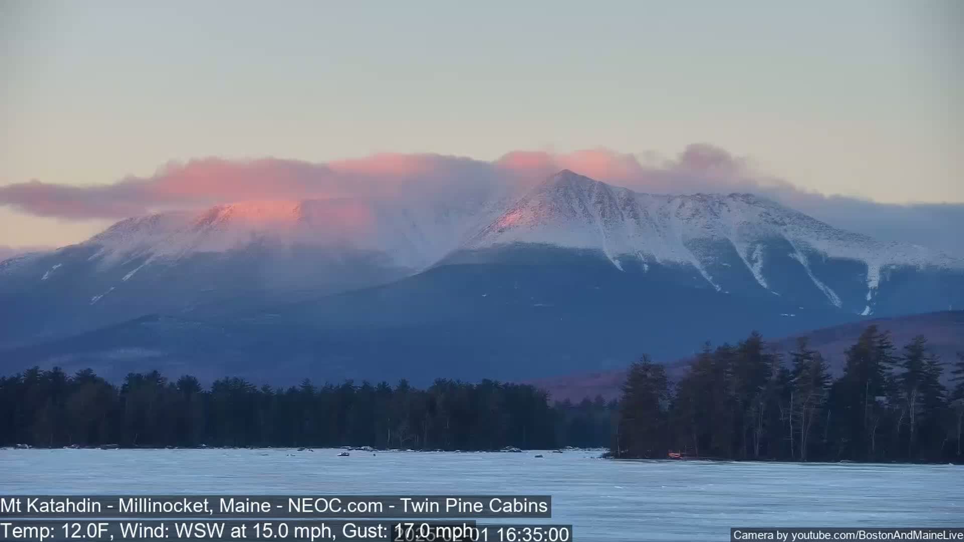 A snow-capped mountain range is bathed in the pink hues of sunset, with a frozen lake in the foreground under a mostly clear sky and a wind blowing from the west-southwest at 15 mph with gusts up to 17 mph.