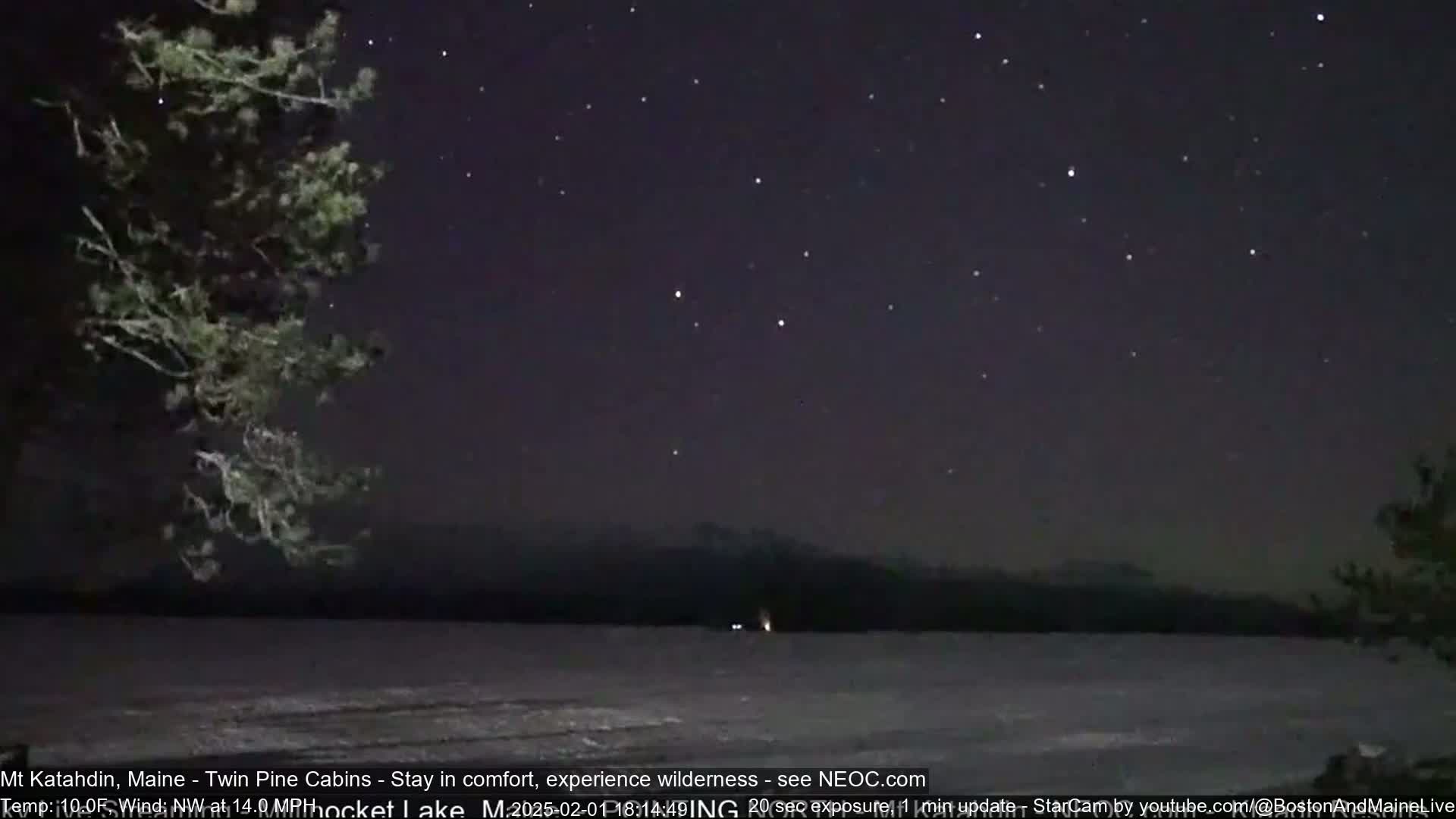 A nighttime view of a calm, snow-covered lake under a clear, starlit sky, with a dark mountain range in the distance and trees on either side of the frame.